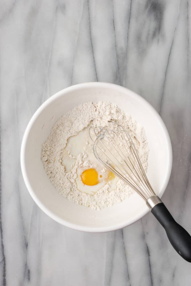 An egg is whisked into flour in a white bowl.