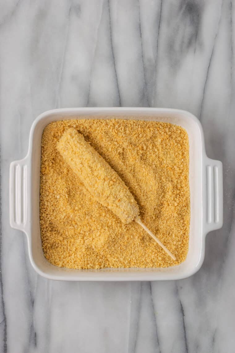 A hot dog on a skewer is rolled in gluten-free panko bread crumbs.