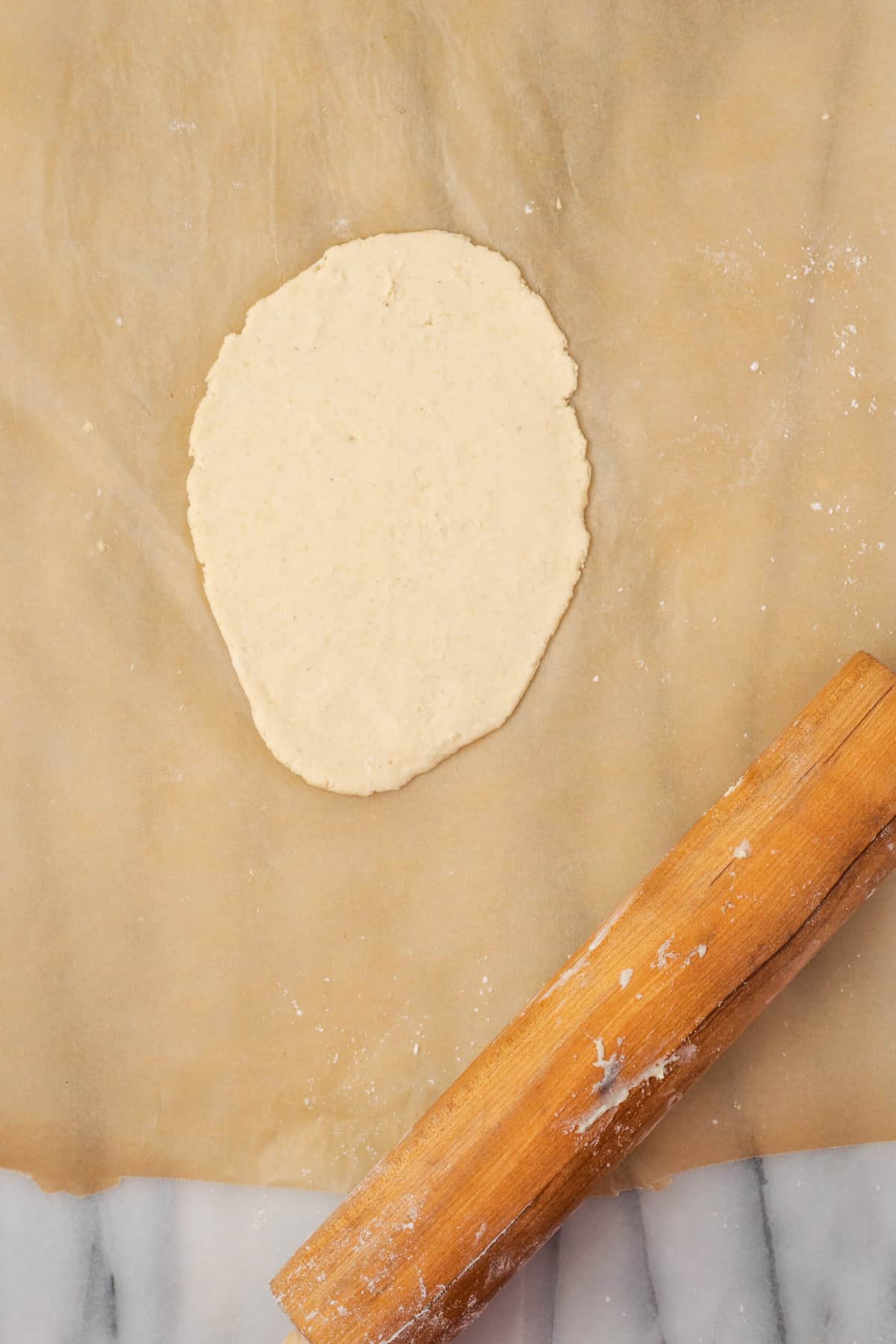 A piece of naan dough is rolled out on parchment paper with a rolling pin.