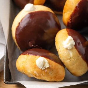 Several bismark donuts with cream filling poking out and chocolate glaze on top in a parchment-lined pan.