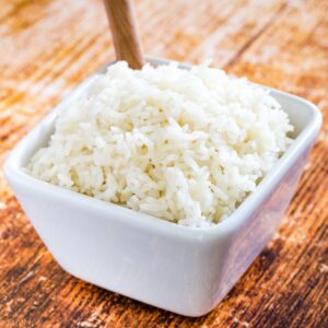 A square white bowl of cooked jasmine rice with a wooden spoon in it.