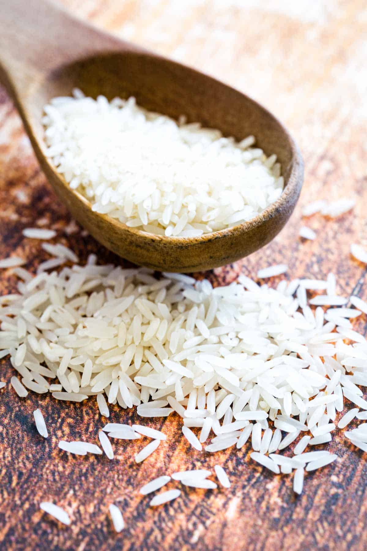 A large wooden spoonful of basmati rice resting on a wooden countertop next to scattered rice grains.