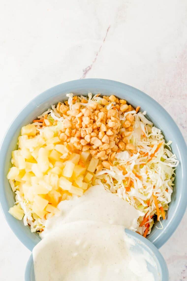 Coleslaw dressing being poured over a large mixing bowl with coleslaw mix, pineapple, and macadamia nuts.