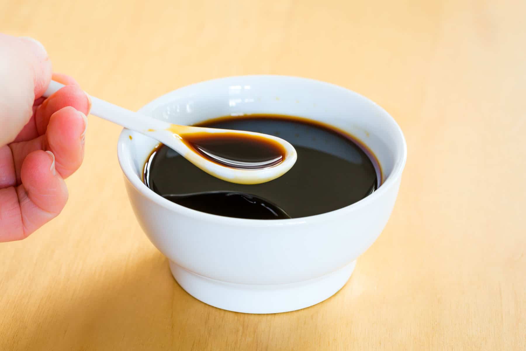 A hand holding a small white spoon of soy sauce over a bowl of soy sauce.