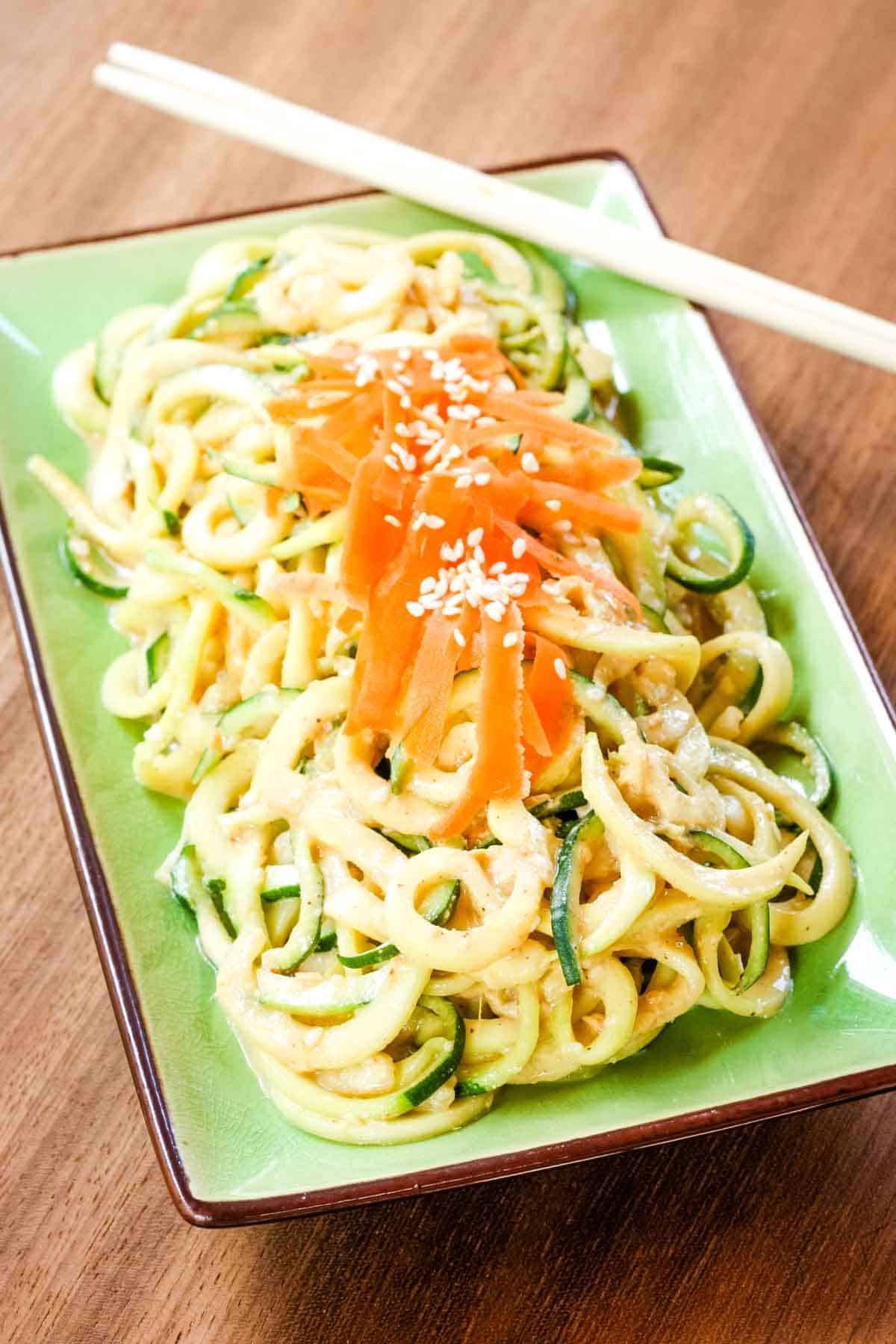 Sesame zoodles garnished with shredded carrots and sesame seed son a green plate with chopsticks on the side.