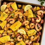 Seasoned shrimp, sausage, corn, and potatoes on a baking sheet garnished with lemon wedges and parsley with text overlay that says "Sheet Pan Shrimp Boil".