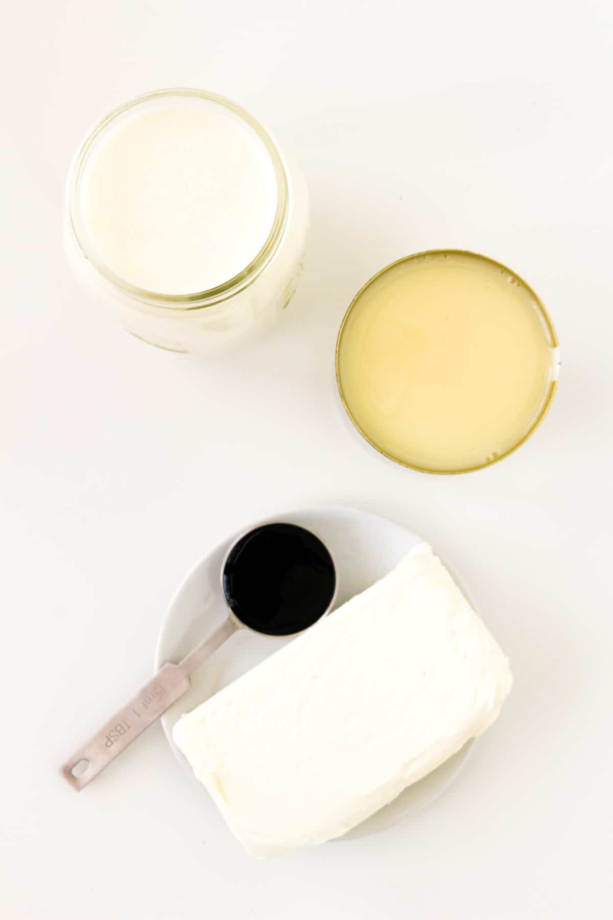 A block of cream cheese, measuring spoon of vanilla bean paste, a can of sweetened condensed milk, and a jar of heavy cream.