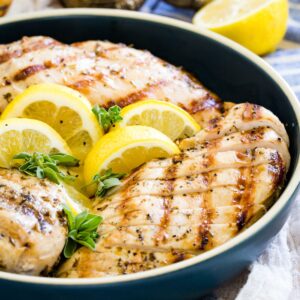 Three grilled chicken breasts with one sliced in a blue dish garnished with lemon wedges and fresh oregano.