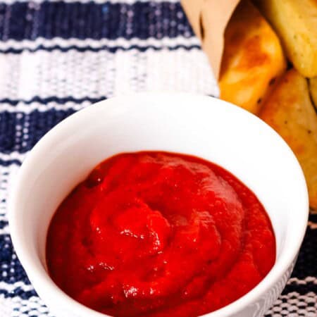 A small bowl of ketchup with fries next to it on a blue and white plaid placemat.