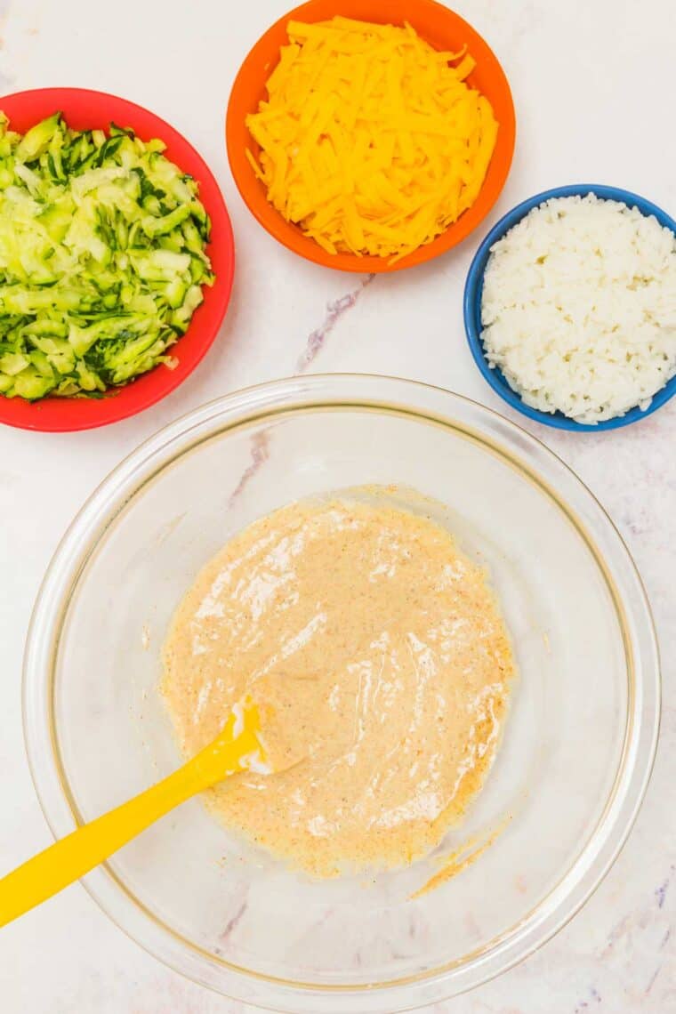 A creamy mixture in a glass bowl surrounded by bowls of shredded zucchini, cooked rice, and shredded cheese.