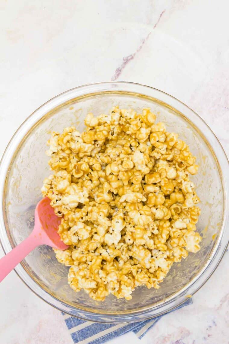A spatula mixes the caramel sauce into the popcorn in a bowl.