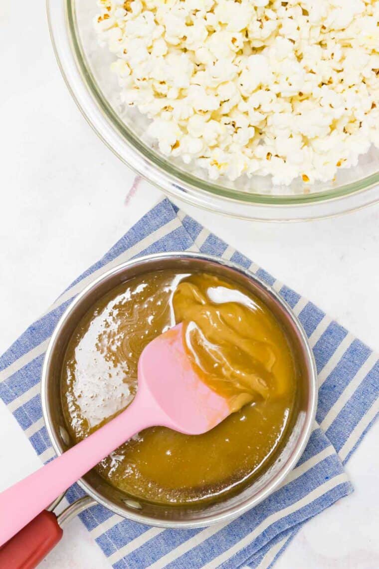 A pink spatula rests in a bowl of caramel sauce with a bowl of popped popcorn on the side.