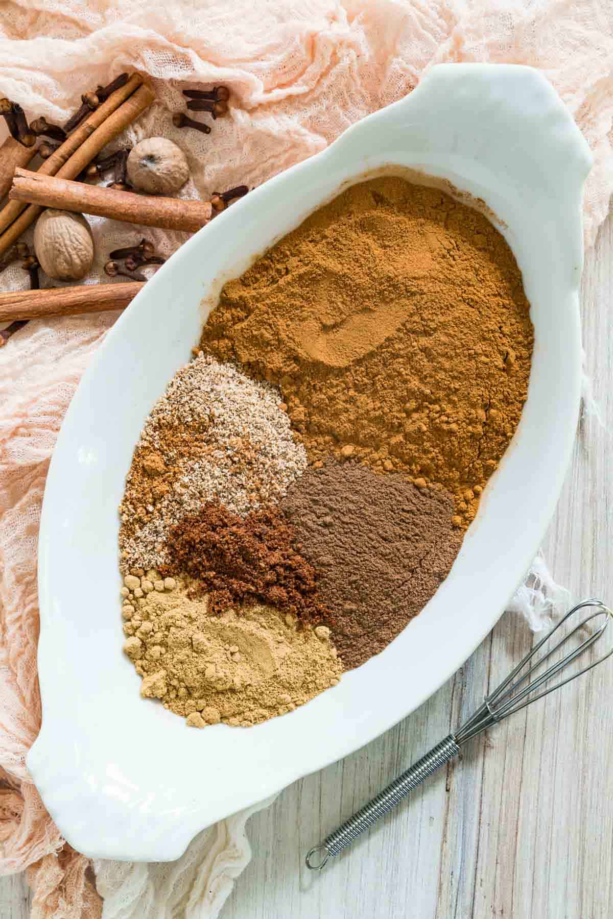Ingredients for pumpkin pie spice mix are shown in a white bowl: ground cinnamon, ginger, nutmeg, cloves, and allspice.