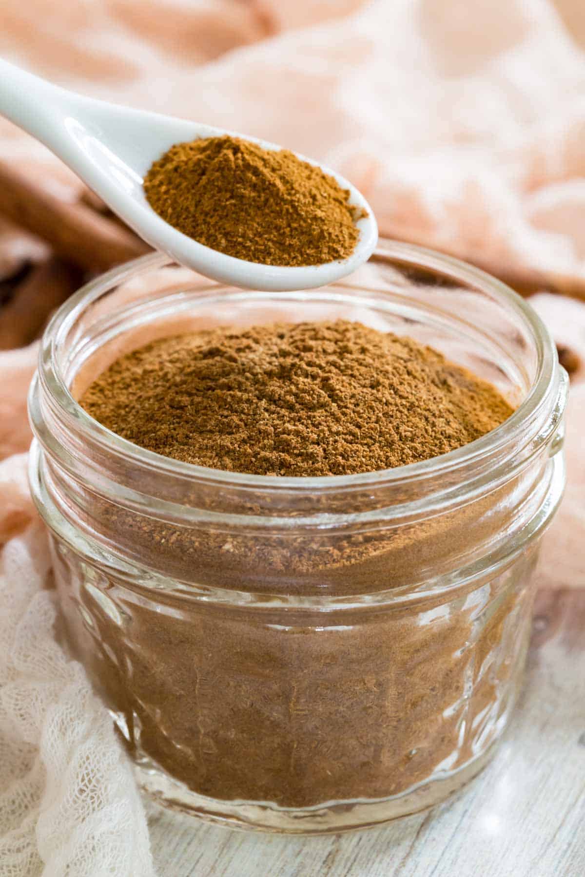 A measuring spoon holds pumpkin pie spice above a jar of spice mix.