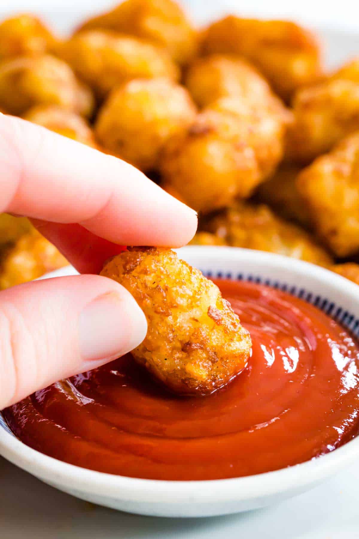 A hand dips an air fried tater tot into a bowl of ketchup.