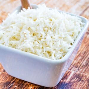 A square white bowl of cooked basmati rice with a wooden spoon in it.