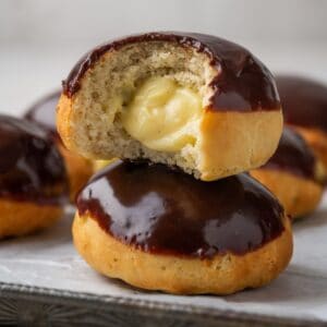 A boston cream donut with a bite out of it on top of another chocolate-glazed donut.