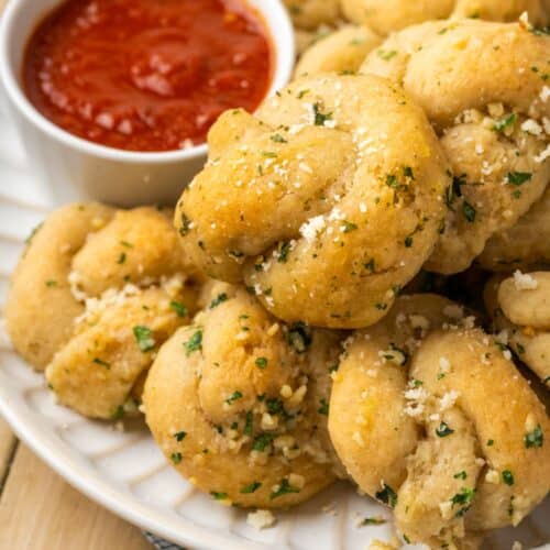A pile of garlic knots on a plate with a small bowl of marinara sauce.