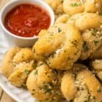 A pile of garlic knots on a plate with a small dish of marinara sauce with text overlay that says "Gluten Free Garlic Knots".