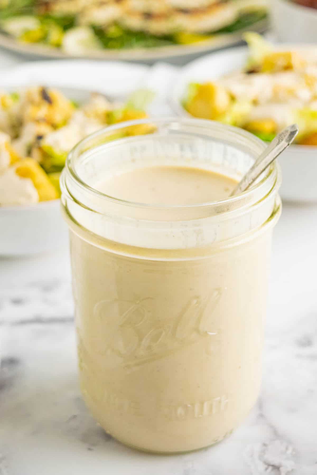 A jar of healthy caesar dressing with a spoon in it and salads blurred in the background.