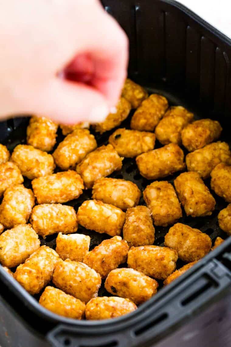 A hand sprinkles salt over tater tots cooking in an air fryer.