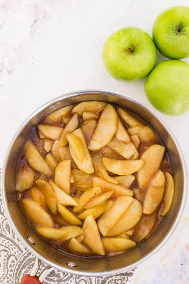 A bowl of spiced apple slices with green whole apples nearby.