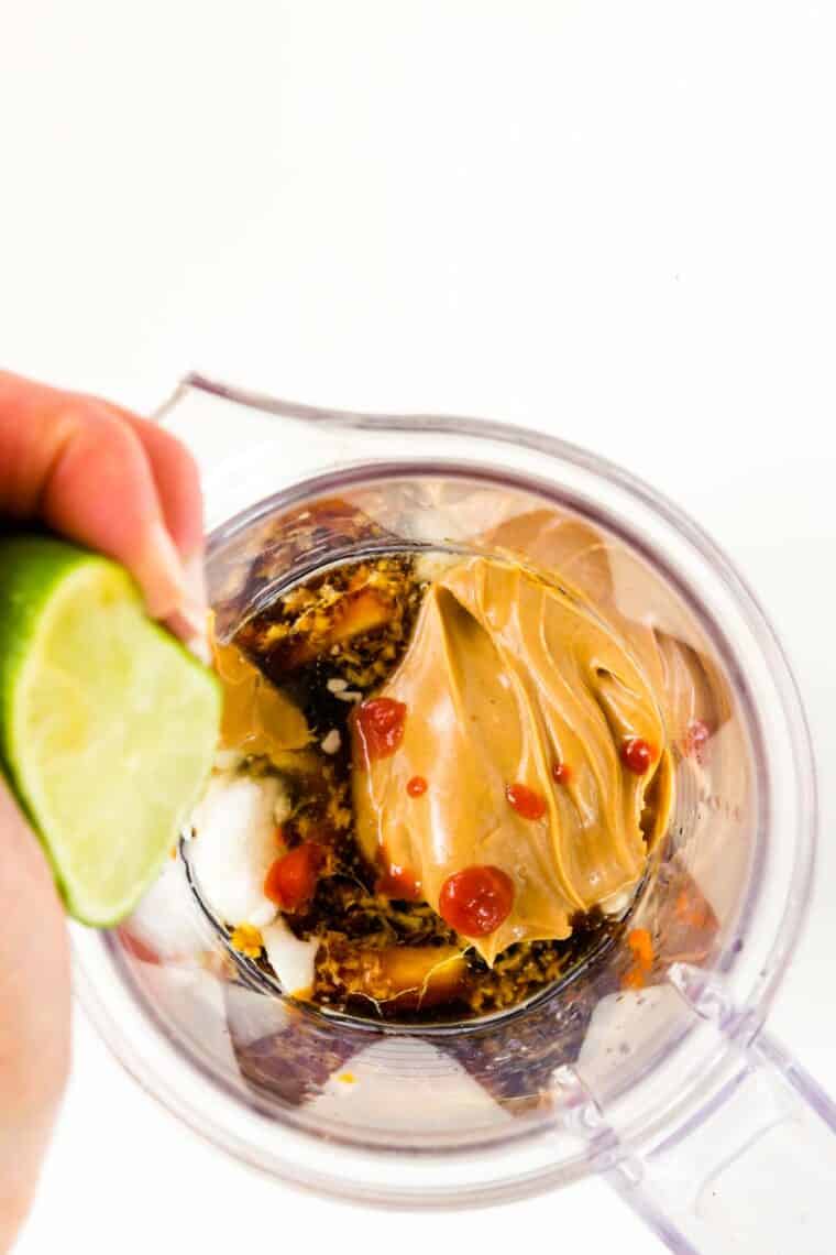 A lime being squeezed over other ingredients including peanut butter and hot sauce in a blender.