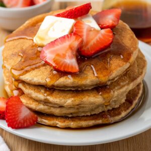 A stack of three oatmeal pancakes topped with pats of butter, sliced strawberries, and maple syrup.
