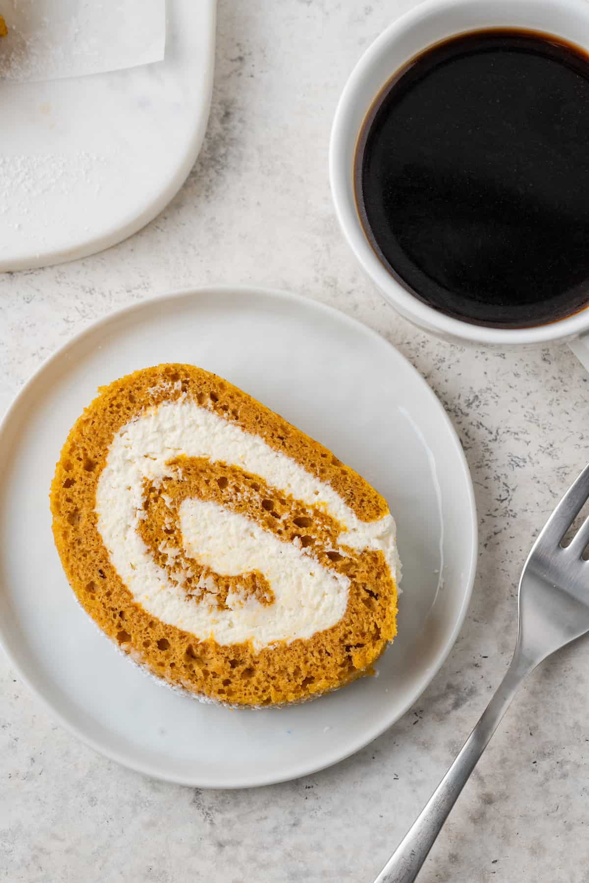 A slice of pumpkin roll on a white plate with a cup of coffee.