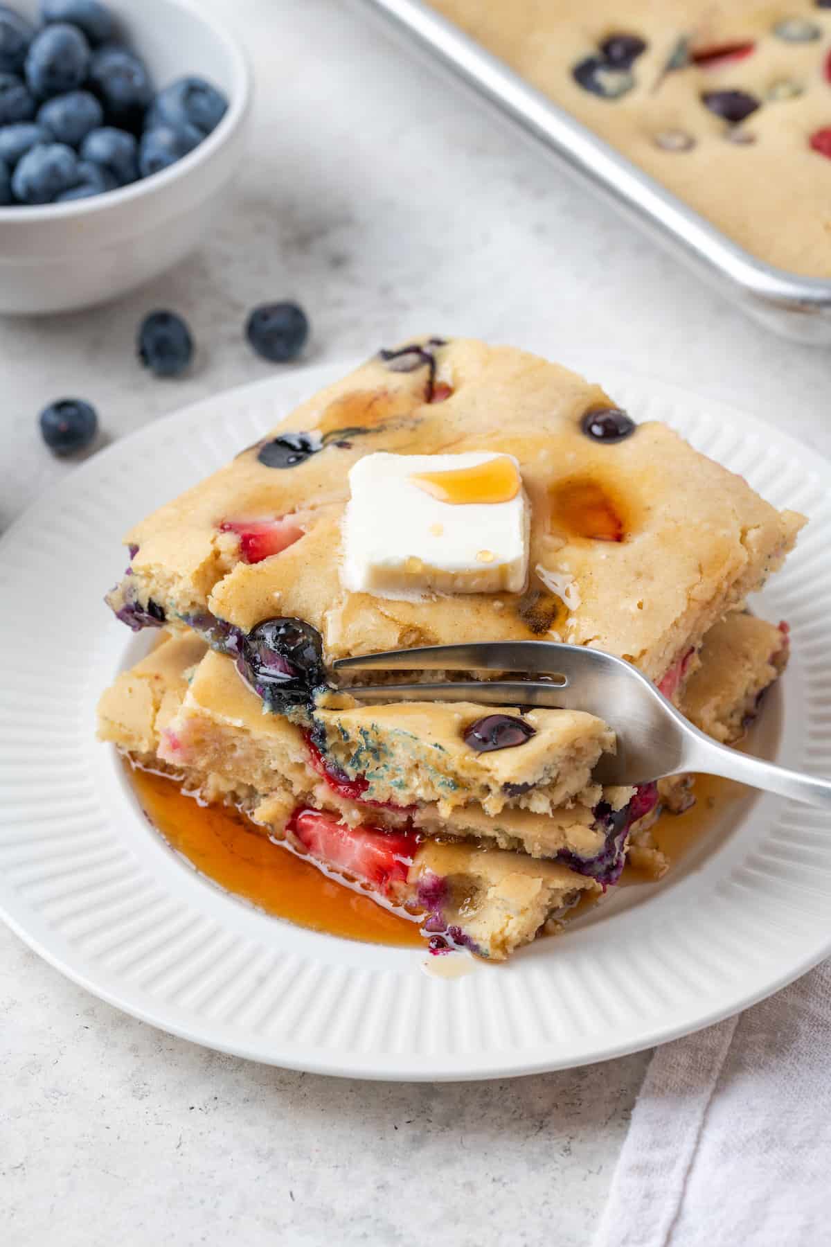 A fork cuts into a plate of berry sheet pan pancakes on a white plate topped with a pat of butter and maple syrup.