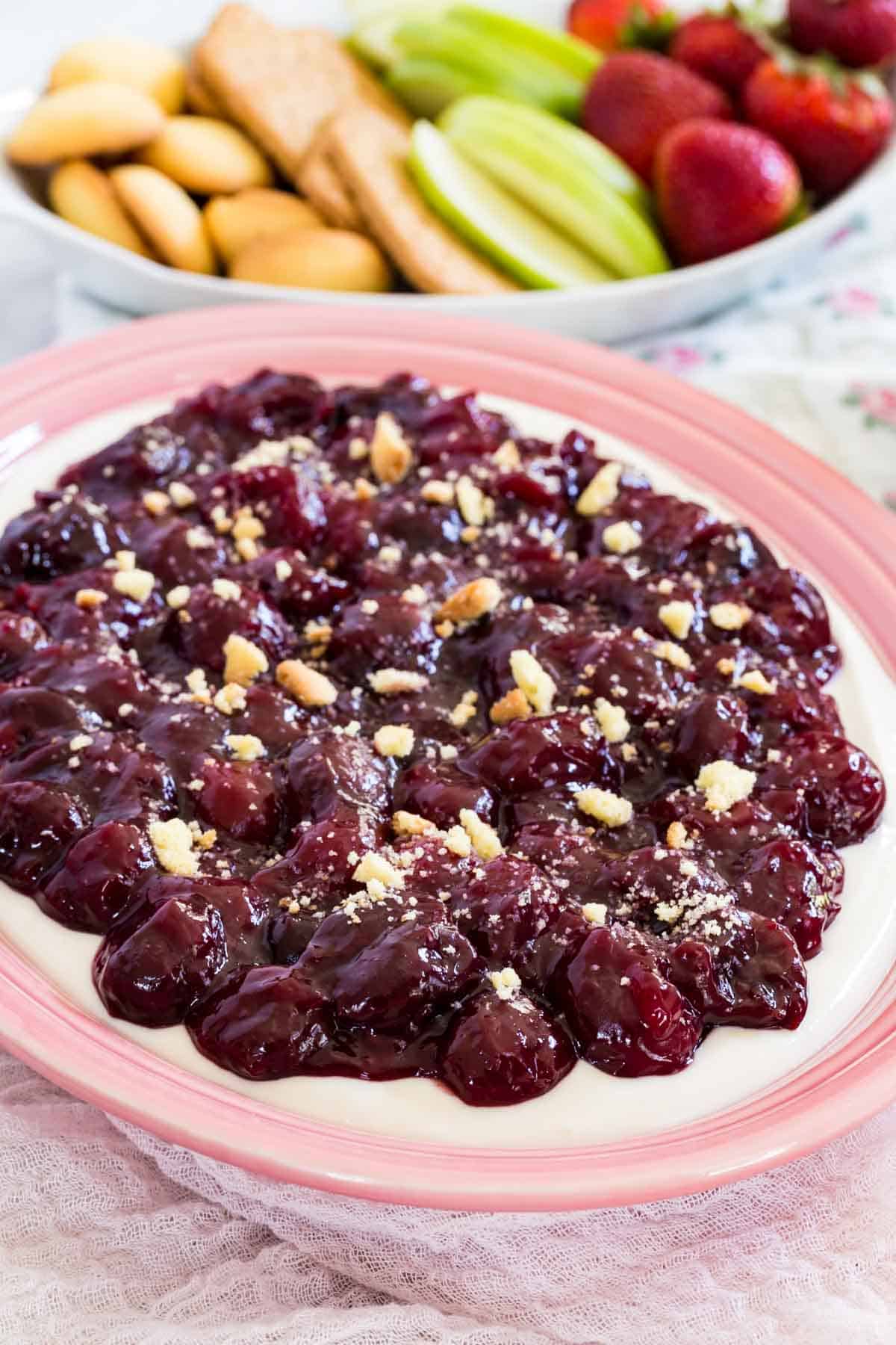 A pink bowl full of cherry cheesecake dip is shown with fruit and cookies in the background.