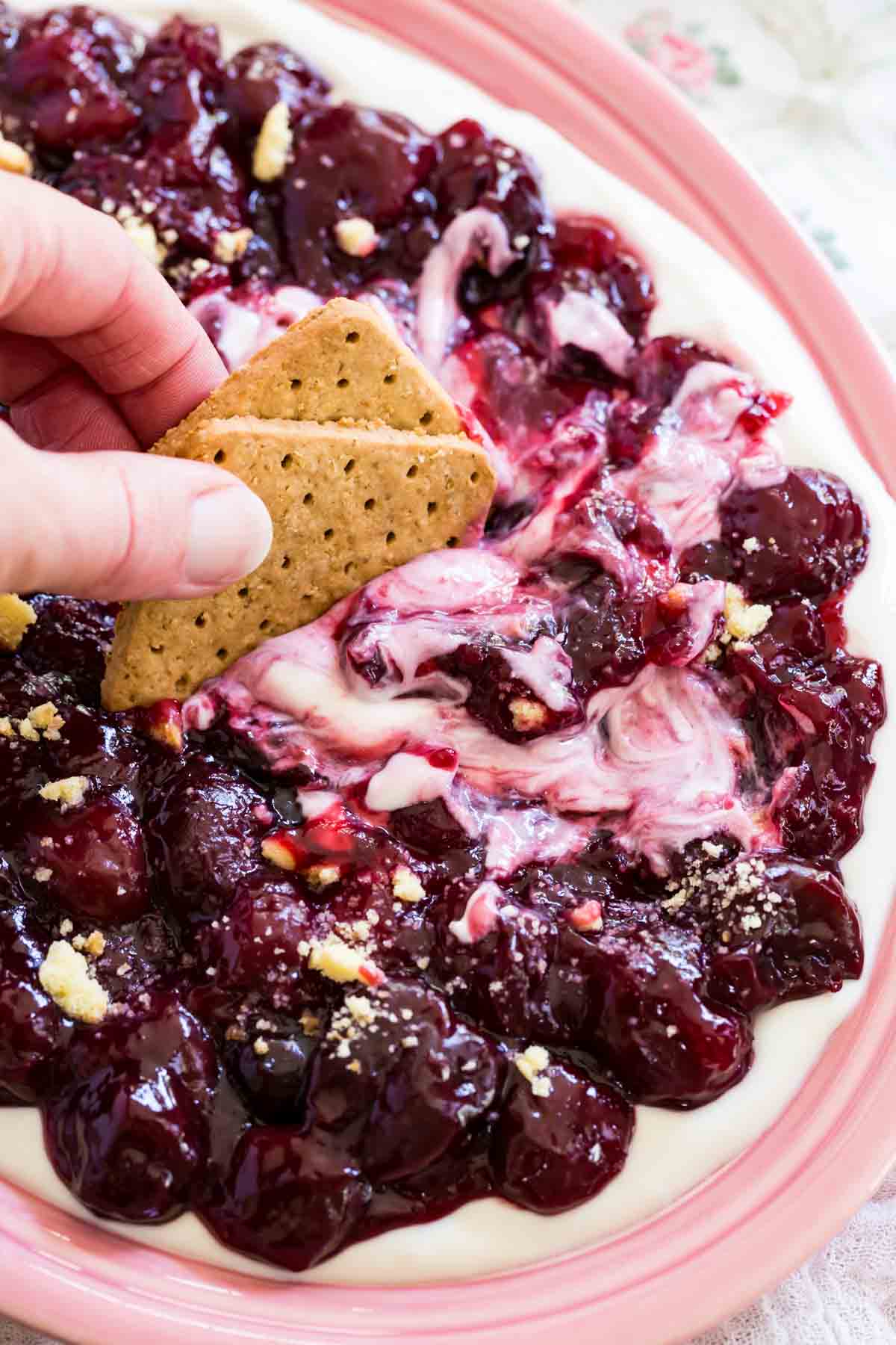 A hand dips a cookie into a bowl of cherry cheesecake dip.