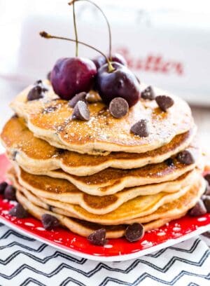 A stack of gluten free chocolate chip pancakes is shown topped with chocolate chips and cherries on a red plate.
