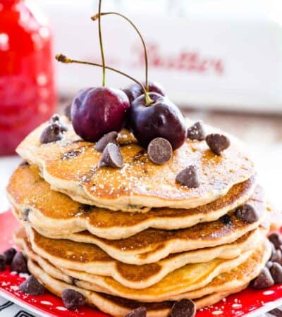 A stack of gluten free chocolate chip pancakes is shown topped with chocolate chips and cherries on a red plate.