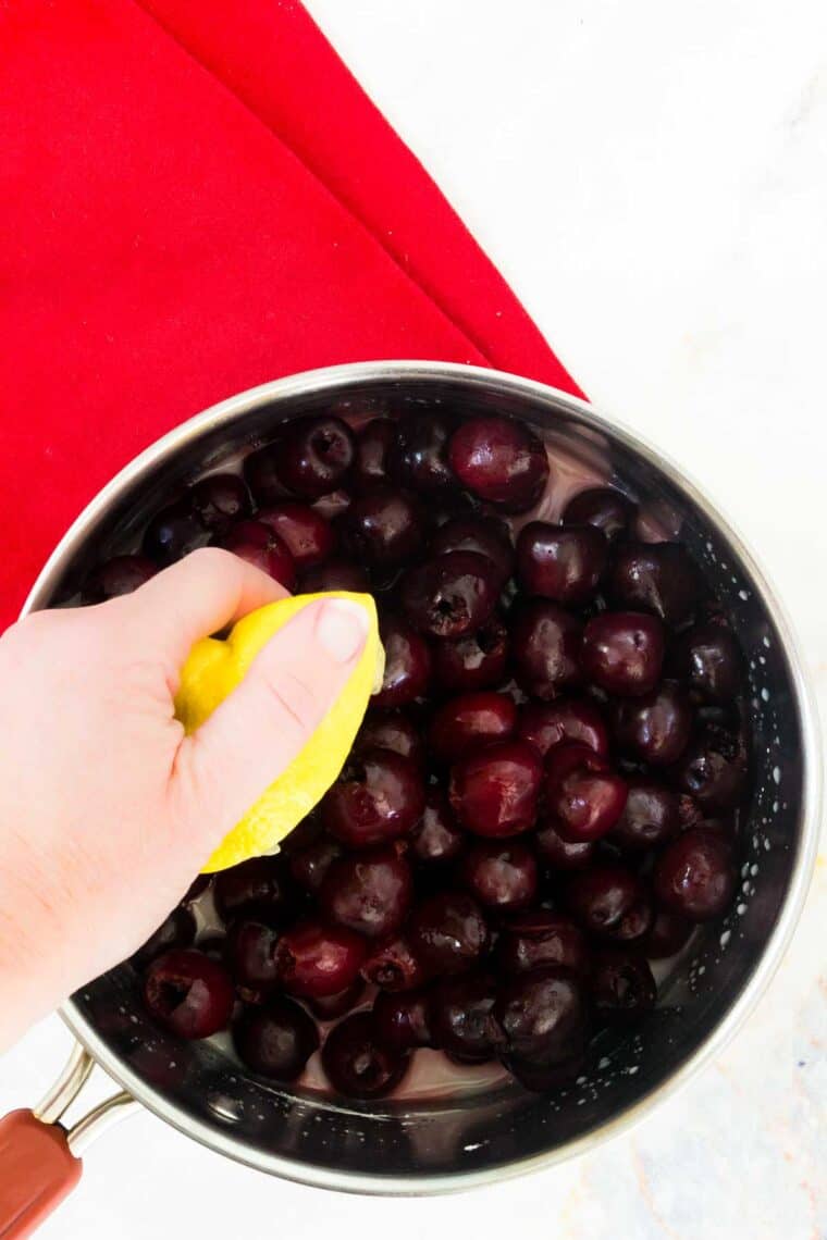 A hand squeezes lemon juice into a pot of cherries.