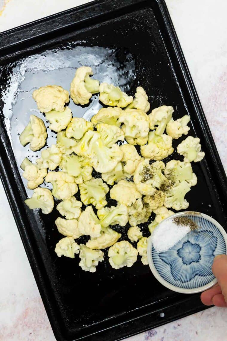 Salt and pepper are added to cauliflower florets on a baking sheet.