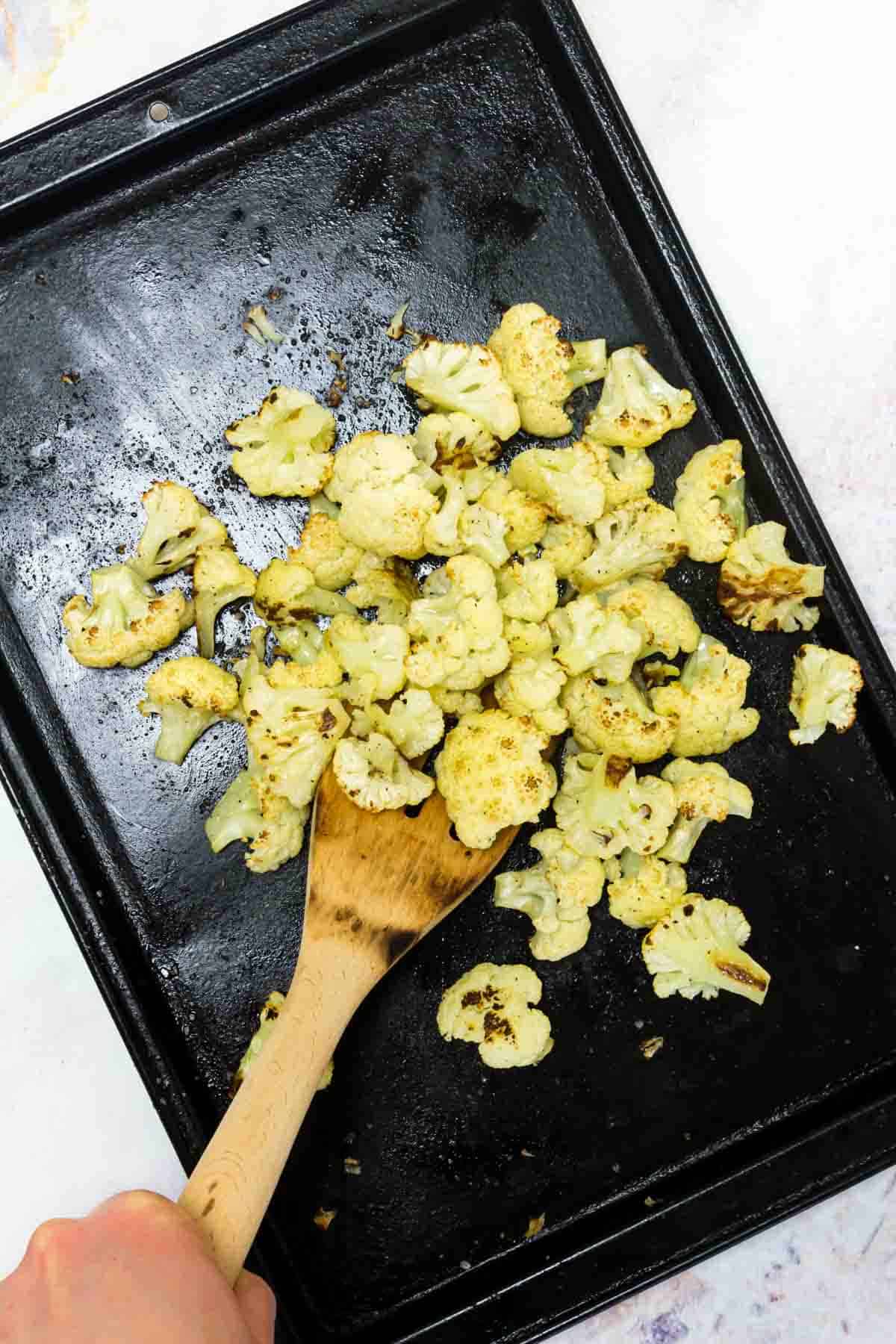 A wooden spoon stirs a baking sheet filled with cauliflower florets.