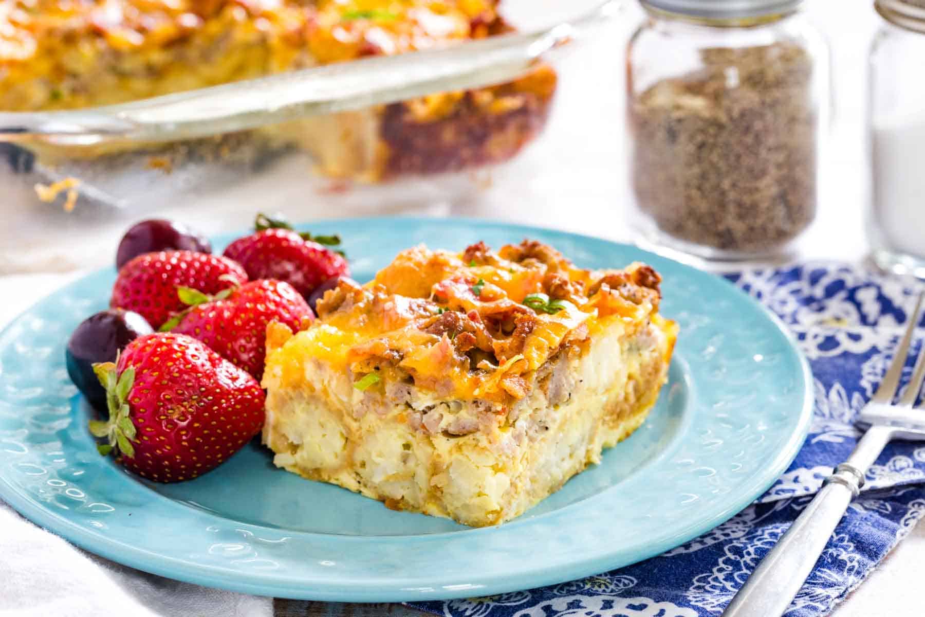 A piece of tater tot breakfast casserole is shown on a blue plate with fresh strawberries.