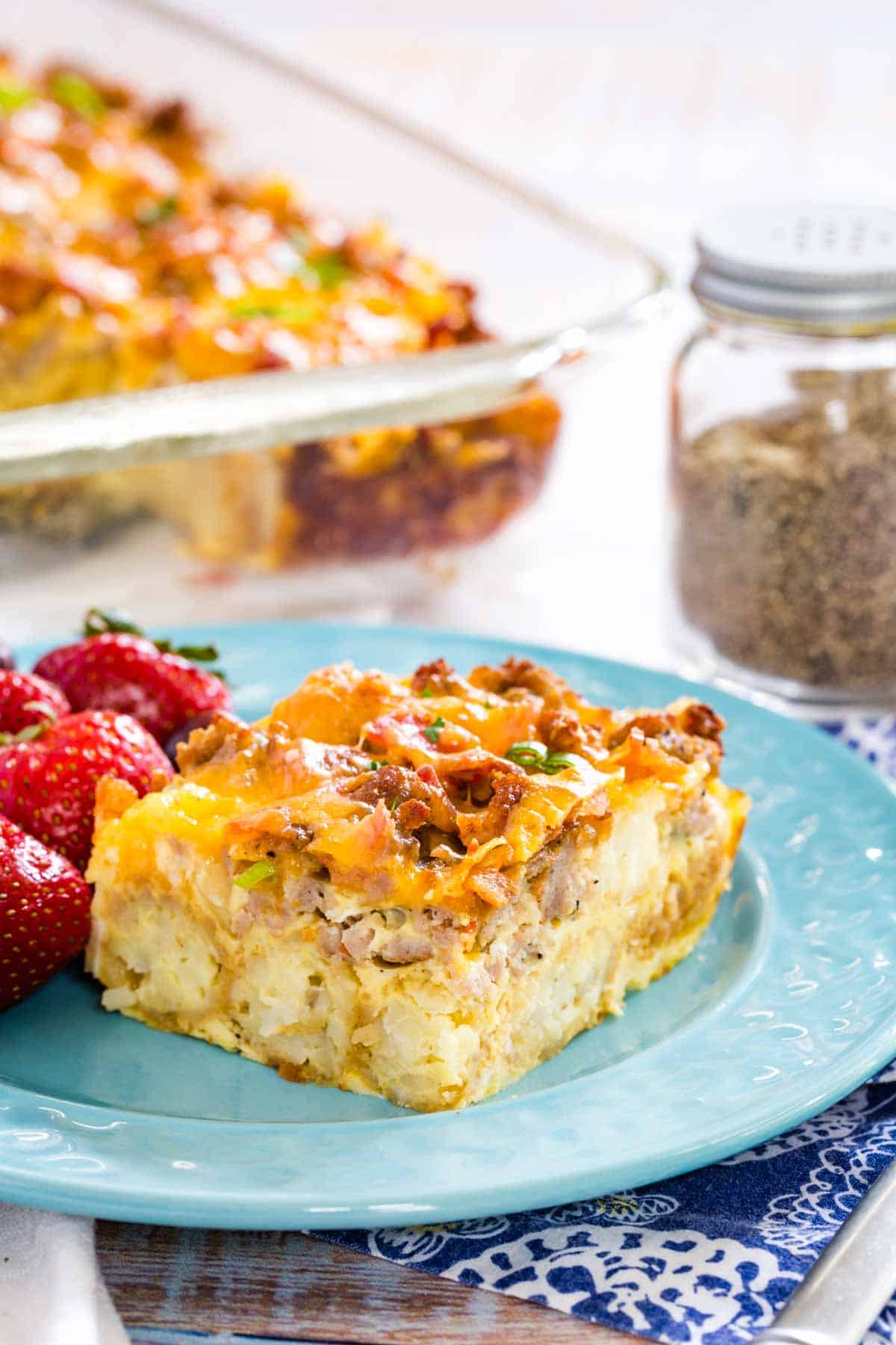 A piece of tater tot breakfast casserole is shown on a blue plate with fresh strawberries.