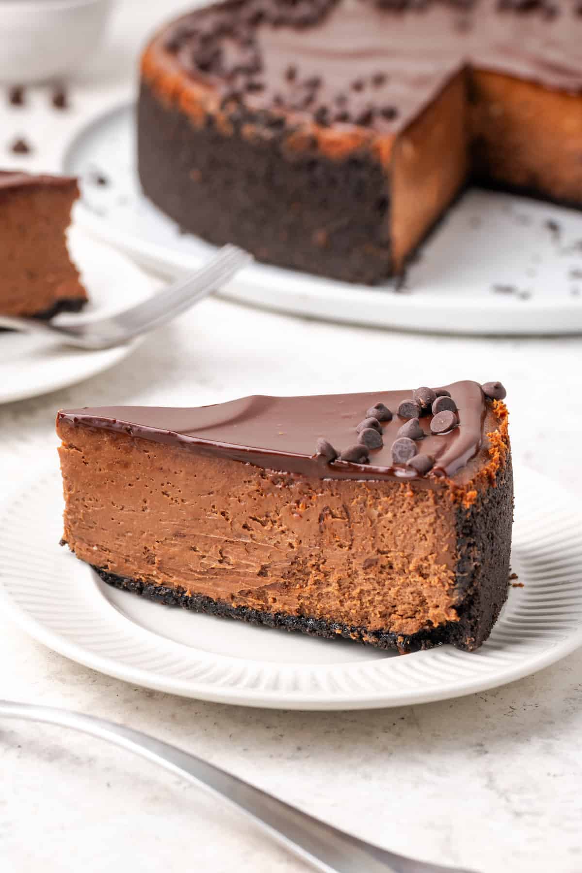 A slice of chocolate cheesecake is shown on a white plate with the cheesecake in the background.
