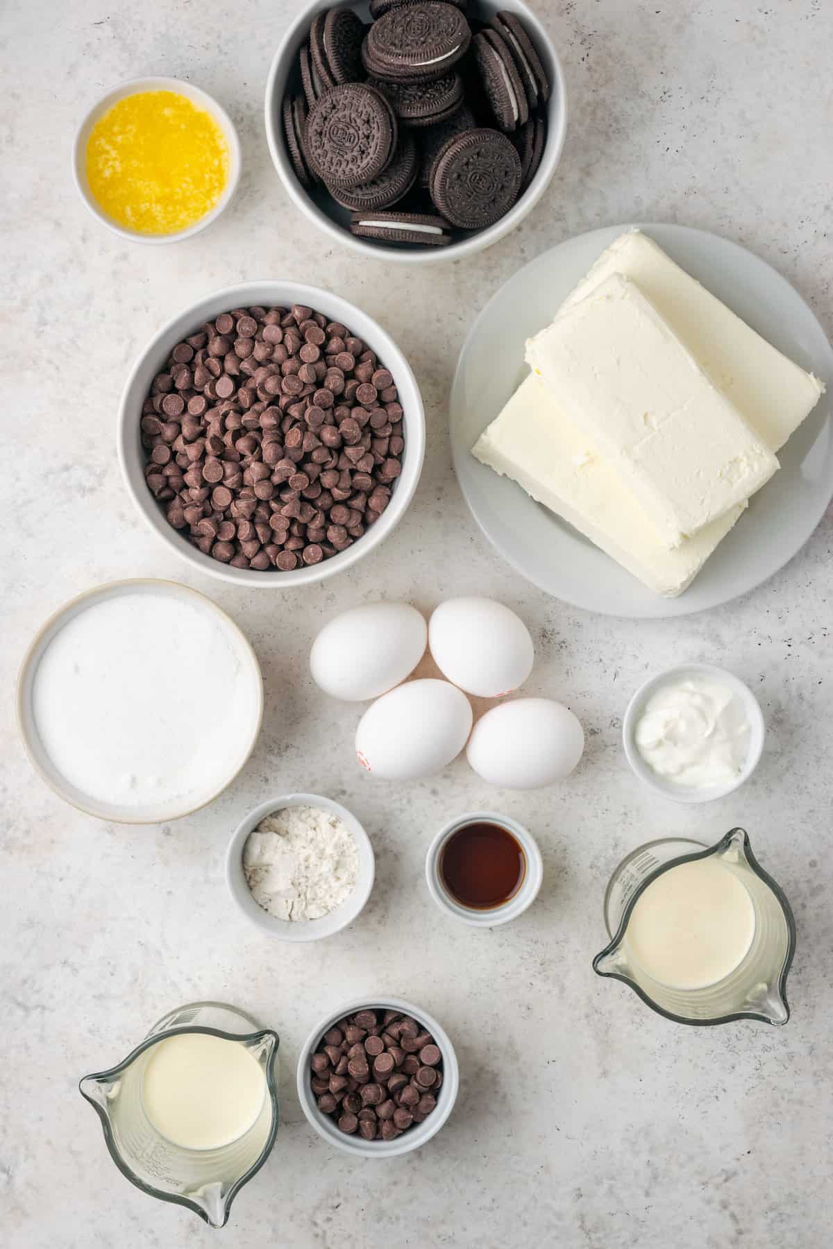 Ingredients needed to make chocolate cheesecake are shown on a white background, including eggs, chocolate chips, cream, cream cheese, vanilla, and gluten free flour.