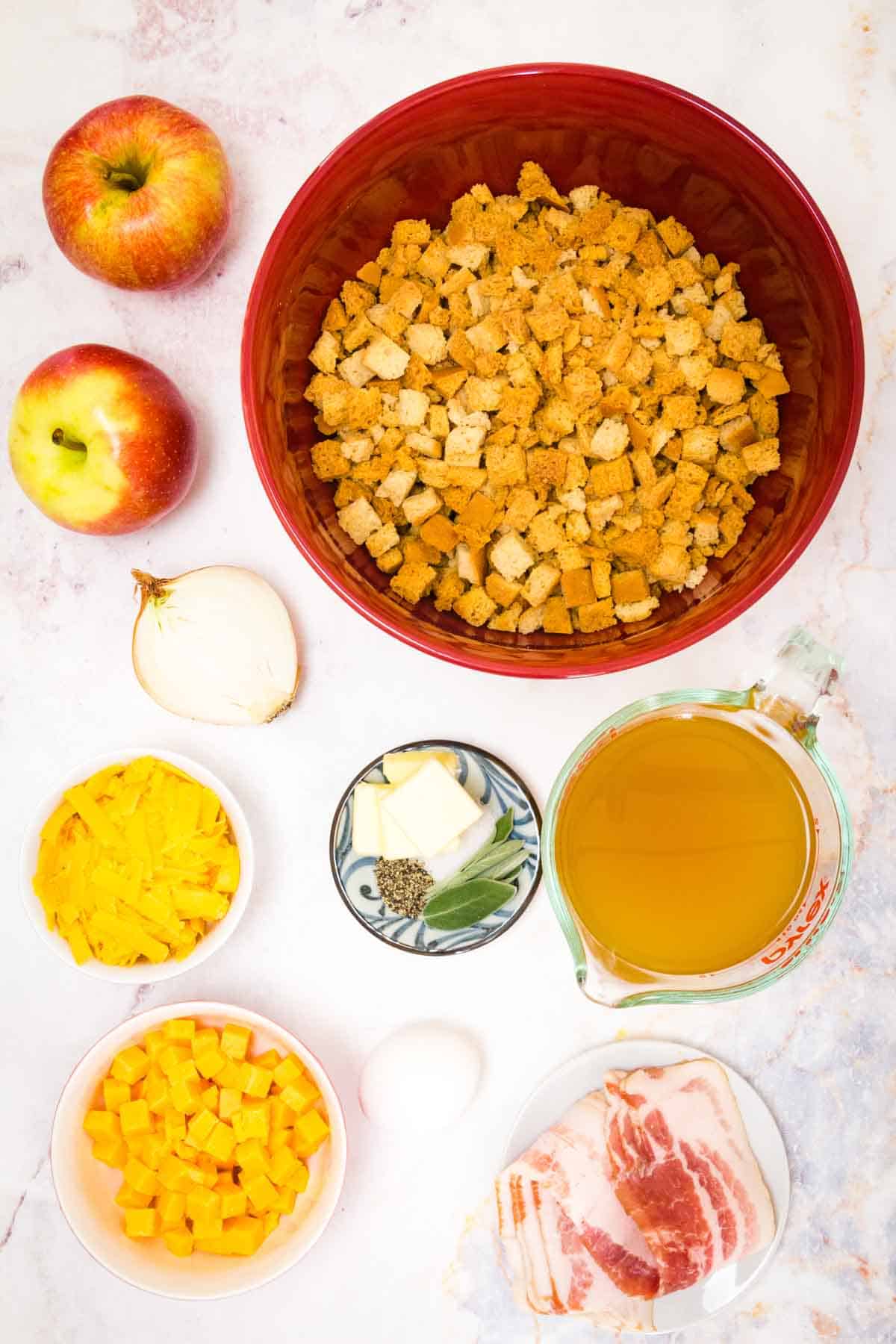 Ingredients for gluten free apple stuffing are shown portioned out: stuffing mix, apples, bacon, broth, cheddar cheese, butter, seasonings.