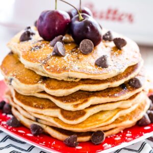 A stack of chocolate chip pancakes topped with cherries and chocolate chips on a red plate.