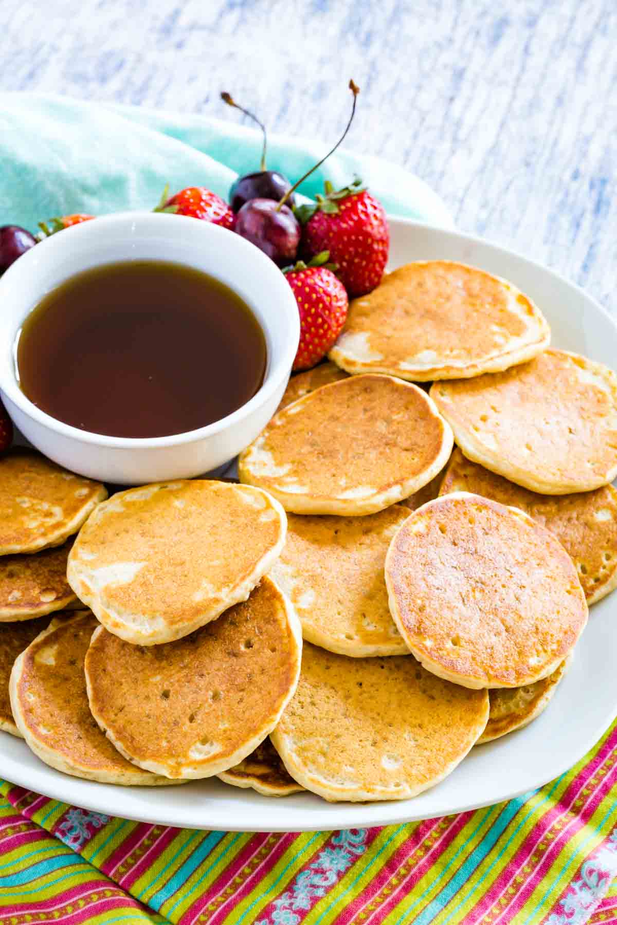 A plate of gluten free silver dollar pancakes with a bowl of maple syrup and strawberries.