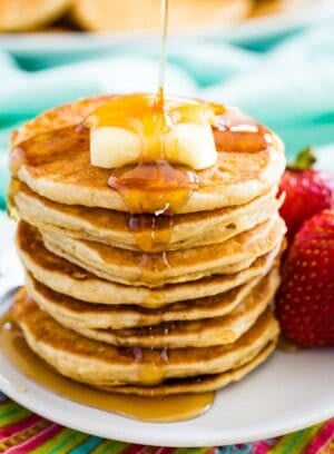 A stack of gluten free silver dollar pancakes topped with butter and syrup is shown on a plate with strawberries next to it.