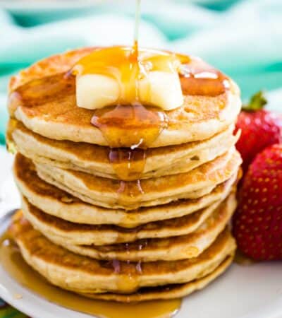 A stack of gluten free silver dollar pancakes topped with butter and syrup is shown on a plate with strawberries next to it.