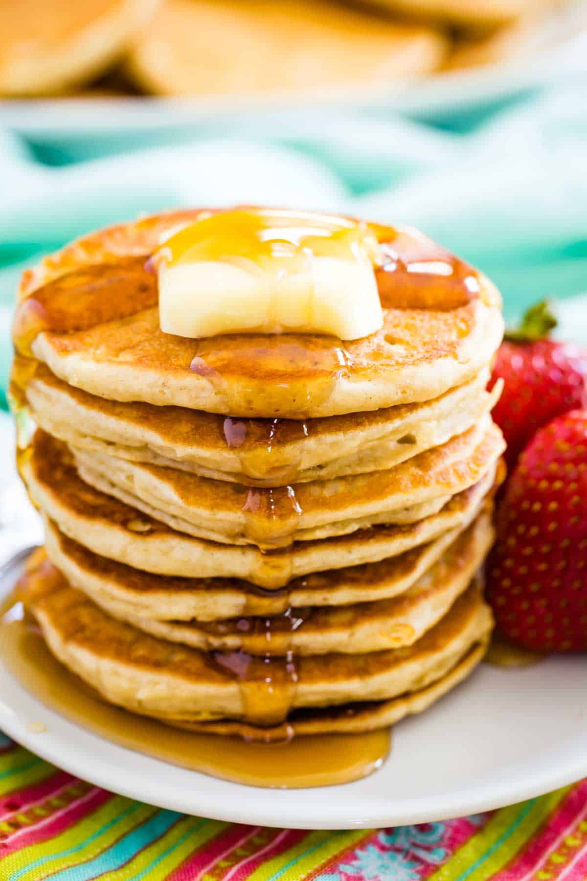 A stack of gluten free silver dollar pancakes topped with butter and syrup is shown on a plate with strawberries next to it.