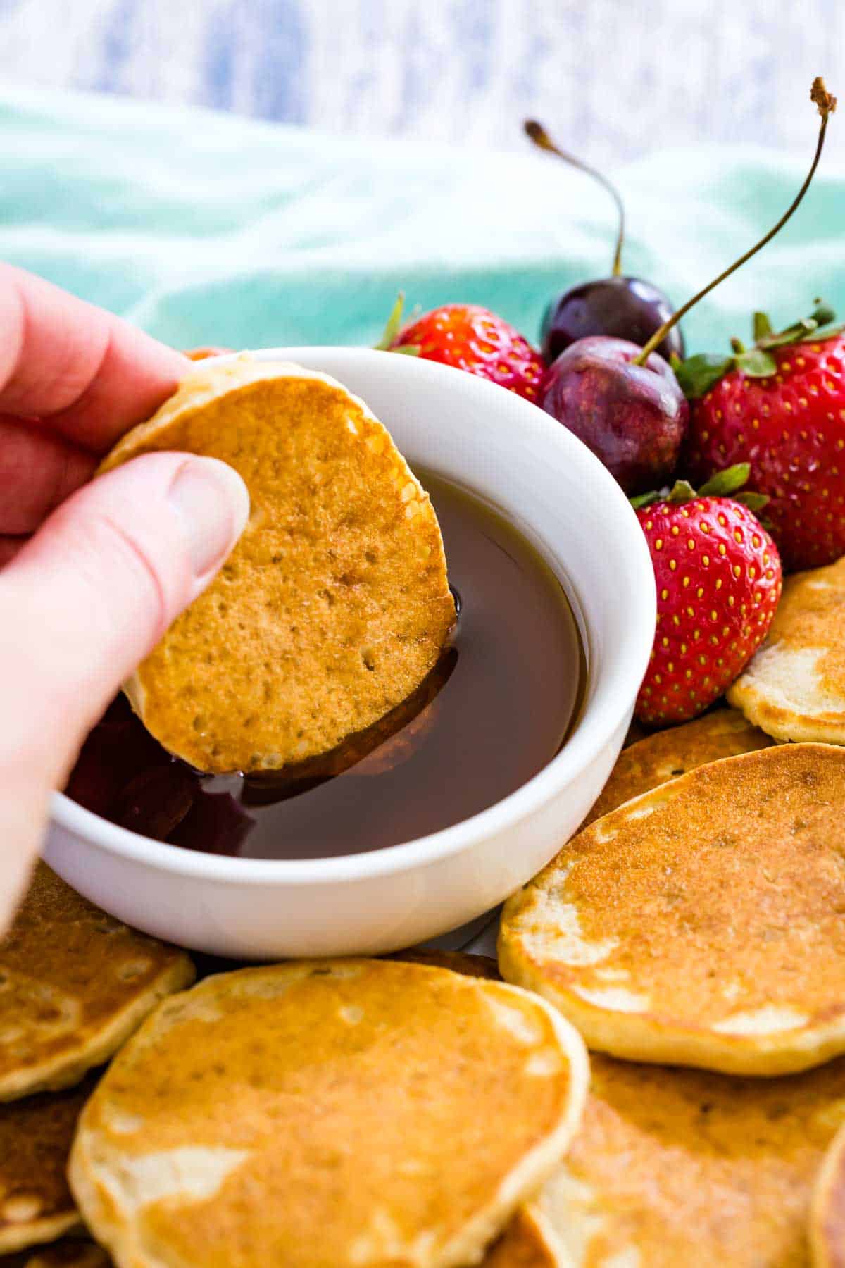 A hand dips a gluten free silver dollar pancake into a bowl of maple syrup.