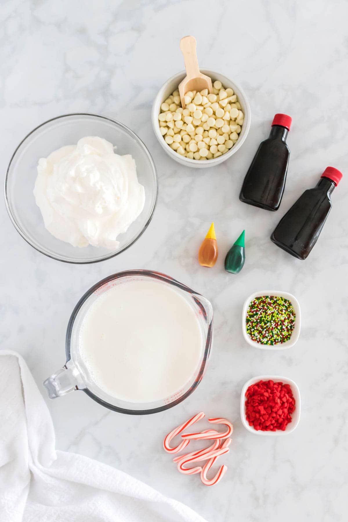 Ingredients to make peppermint hot chocolate are shown on a white background: vanilla, peppermint extract, white chocolate chips, milk, whipped cream, food coloring, sprinkles, and candy canes.