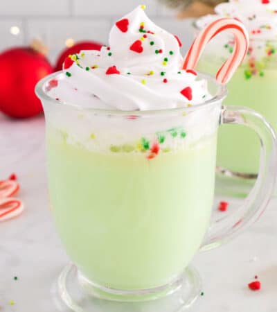A green glass of peppermint hot chocolate topped with whipped cream, sprinkles, and a candy cane.